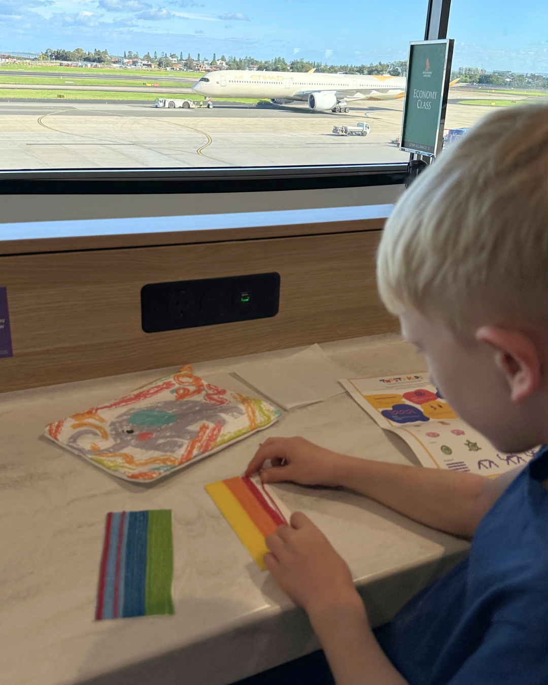 A young child is sitting at a table by a large airport window. The child is interacting with colorful wax sticks on the table, with printed cards nearby.