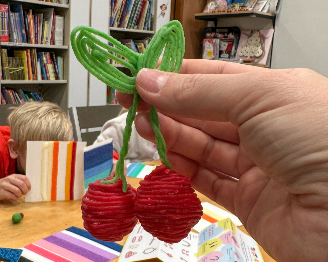 A hand holding 3D cherries made with red and green wax sticks at the Twisty Kids workshop