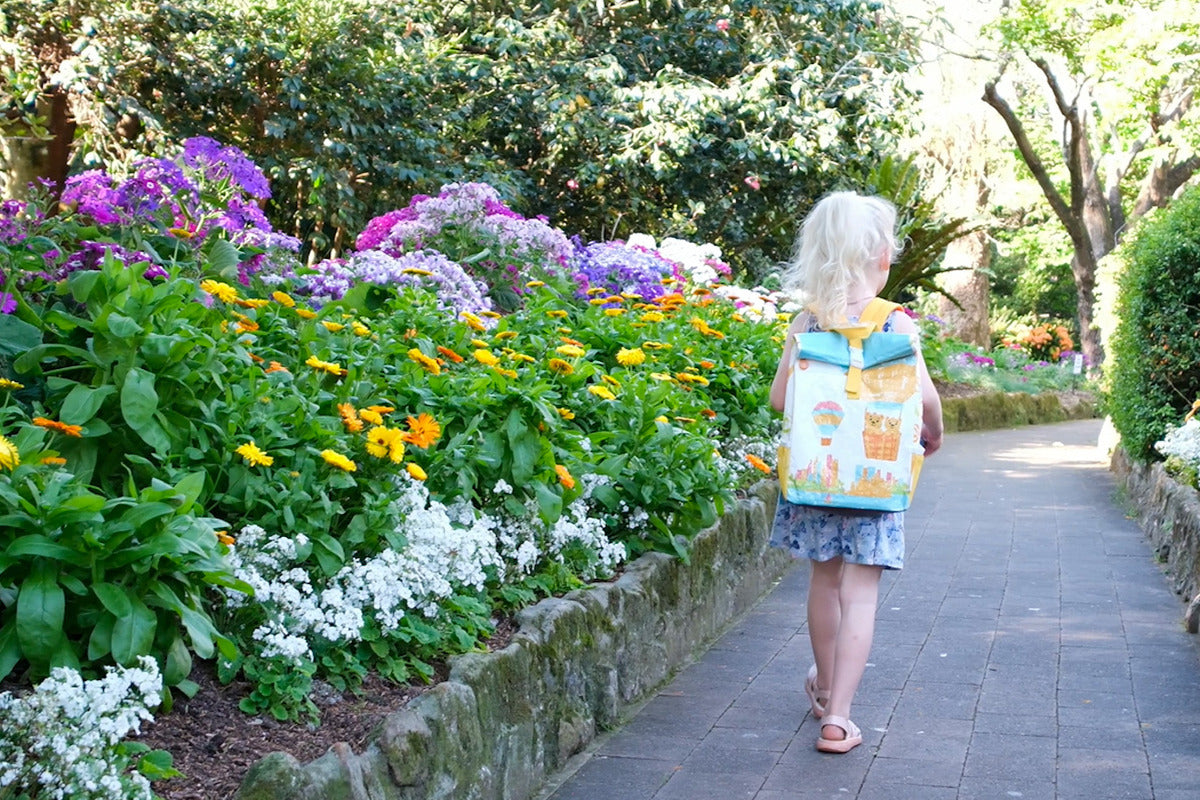 A young child with blonde hair walks along a flower-lined path wearing a colourful travel backpack. 