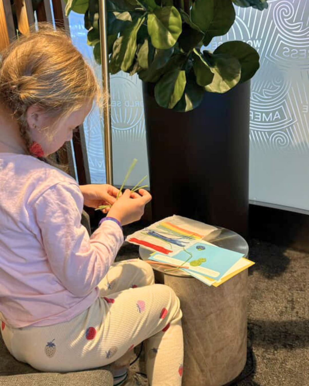 A child sits on a bench focused on a toddler activity, shaping colourful reusable wax sticks on a small round table.