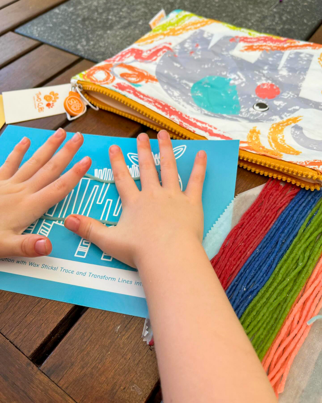 A child's hands are pressing down on a blue printable card with wax sticks on a wooden table, with a zippered washable pouch.