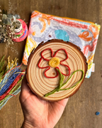 A hand holds a round wooden slice decorated with a colorful embroidered flower in red, yellow, and green yarn. A patterned zip pouch and other craft items are visible in the background on a wooden surface.