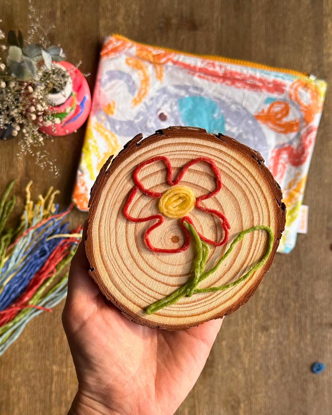 A hand holds a round wooden slice decorated with a colorful embroidered flower in red, yellow, and green yarn. A patterned zip pouch and other craft items are visible in the background on a wooden surface.