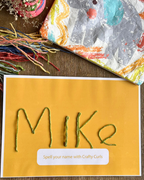 A craft card featuring the name 'Mike' spelled out in green yarn curls on a mustard yellow background, with the text 'Spell your name. The card is styled on a wooden surface alongside colorful yarn strands and a patterned fabric.
