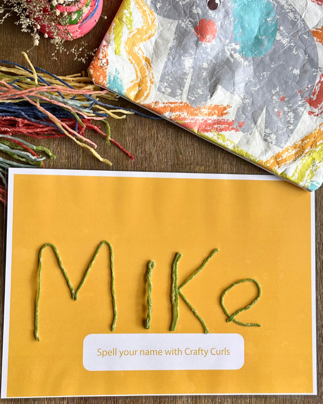 A craft card featuring the name 'Mike' spelled out in green yarn curls on a mustard yellow background, with the text 'Spell your name. The card is styled on a wooden surface alongside colorful yarn strands and a patterned fabric.