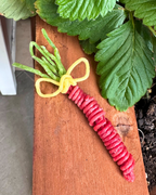 A carrot-shaped decorative item made from red spiral pasta, with green curly pasta stems and a yellow curly pasta bow, placed on a wooden surface with leafy greens nearby.